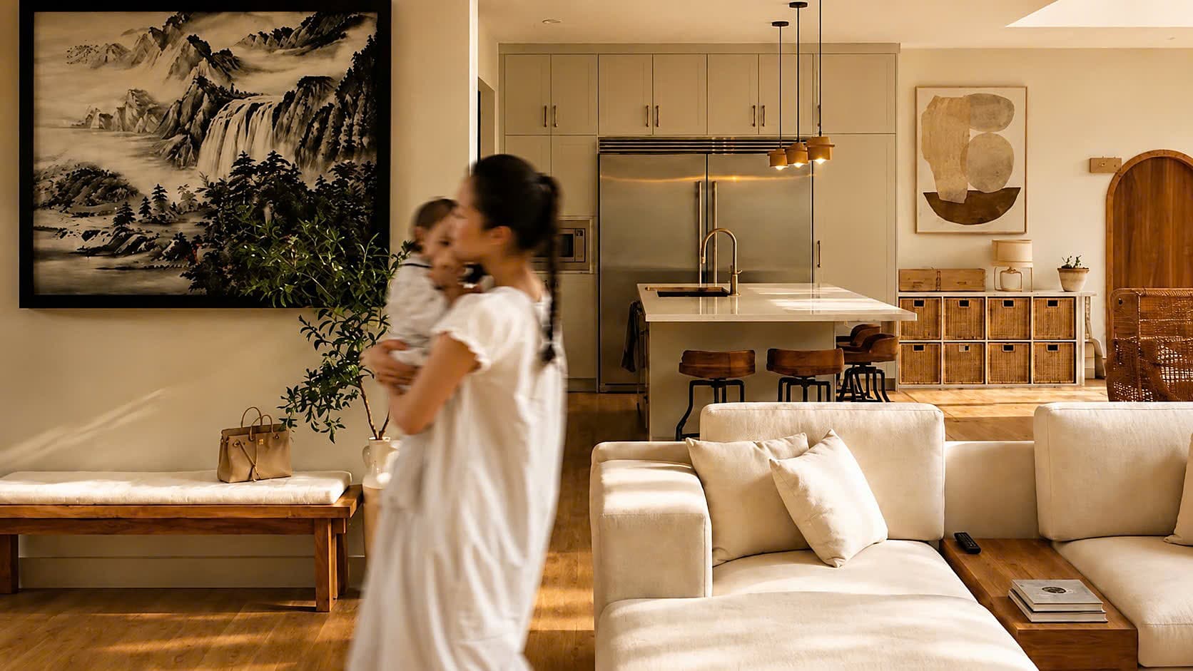A mother and child in a warm, sunlit open-plan living and kitchen space with natural wood and cream furnishings